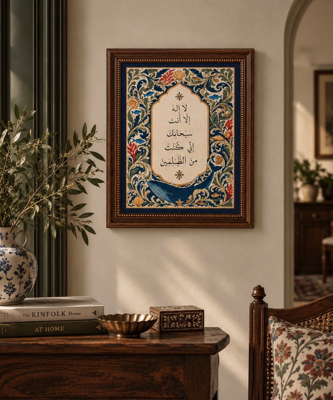 A framed Islamic wall art print featuring Arabic calligraphy and an ornate floral border, displayed on a beige wall in a styled room with a wooden console table, books, and a vase.